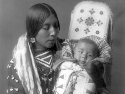 A Crow woman holding an infant in a decorated cradleboard, photograph by Edward S. Curtis, c. 1908.