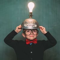 A young boy dressed in retro 1980s attire, with bow tie and eyeglasses, wears a light bulb idea invention machine to help him think of the next big idea. (nerd, nerdy, thinker) SEE CONTENT NOTES.