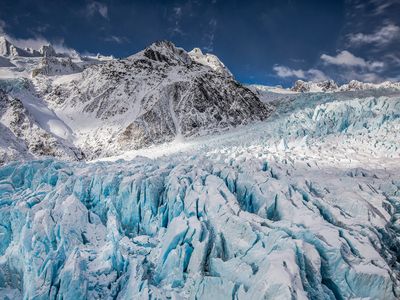 A gray mountain stands behind a white and blue jagged glacier. Above is blue sky with a few clouds.