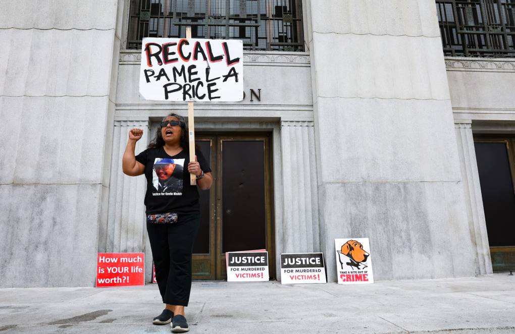 A woman stands in front of an official-looking building holding a sign that says 'Recall Pamela Price.'