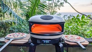 Gozney Arc XL pizza oven fired up on its stand with two uncooked pizzas on peels on fold-up shelves