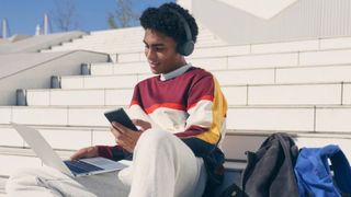 Back to school headphones, student sitting on stairs wearing headphones while typing on a laptop and holding a phone