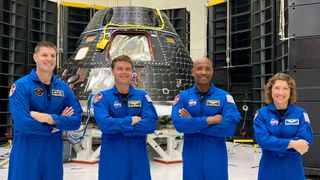 four astronauts standing in a row with arms crossed. they wear flight suits. behind them is a cone-shaped spacecraft