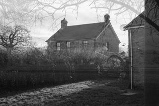 A black and white double exposure of a farmhouse with the silhouette of trees around the edges of the frame