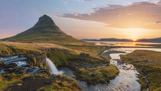 a tall mountain in the background looks like a witches hat and there is a raging waterfall in the foreground. A orange yellow can be seen just above the horizon.