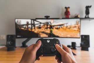 A person holding an Xbox controller in the foreground. The background, which is blurred, shows a game on a wide-screen TV