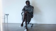 Man sits in chair in Moncler grey puffer jacket in concrete room with white wall