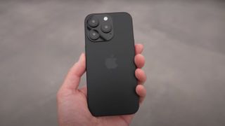 A black iPhone 16 Pro dummy model held in the hand with its back to the camera so the Apple logo is visible.