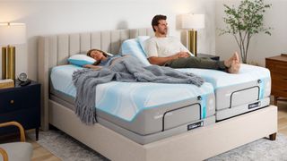 A couple on the Tempur-Pedic Tempur-ActiveBreeze smart bed. The man sits up with the head of the bed raised while the woman lays down sleeping next to him under grey blanket in chic bedroom with gold and white lamp, suede bed frame and a plant. 