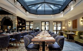 A restaurant with rows of decorated dining tables, a patterned rug, a wall mirror, wall shelving under a large skylight.