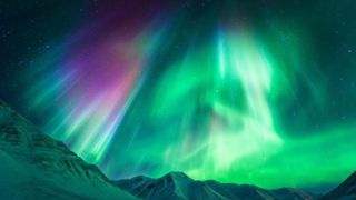 Ribbons of green and purple light dance across the night sky above snow-capped mountains.