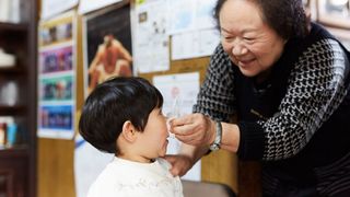 a grandmother smiles as she pinches a tissue over her young grandchild's nose; both are standing in what looks like a store with posters on the walls