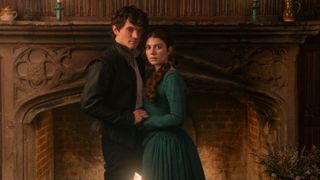 Guildford Dudley (Edward Bluemel) and Lady Jane Grey (Emily Bader) holding hands by a fireplace in "My Lady Jane"