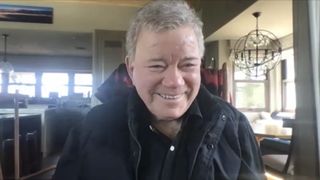 a silver-haired man smiling, wearing a black button-up shirt and black coat.