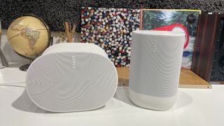 The Sonos Era 300 and Sonos Move 2 in white side by side on a white surface with a globe and vinyl album covers in the background.