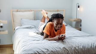 A woman wearing an orange top laying on her stomach on bed in light blue room as she smiles at her phone.