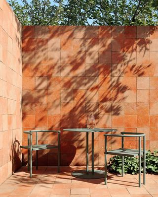 Outdoor green matching chair (2) and round table with a jug of water placed on the table, set outdoor in a courtyard with matching two-toned brown walls and floors with square bricks and green trees in the backdrop