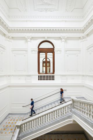 Two people walking down a white decorated staircase with a large window and a decorated roof above it.