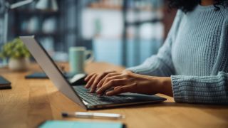 A close up of a person sitting at a table and typing on a computer (a laptop)