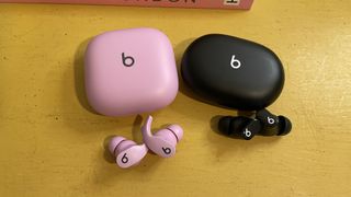 A pair of pink Beats Fit Pro and black Beats Studio Buds with their charging cases, lying on a yellow surface.