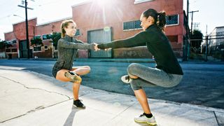 Two women holding hands performing a standing pigeon pose with left foot over right knee. Outside on pathway