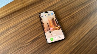 Apple iPhone 15 Pro Max lying on a wooden table. The homescreen features a wallpaper of a city street scene.