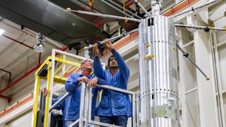 Two people on a scaffold tend to the protruding arms of metallic cylinder 