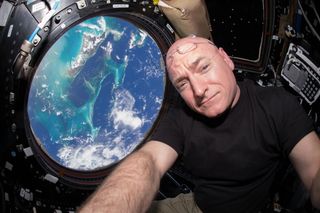 a man in a black t shirt poses in front of a window, through which earth can be seen far below