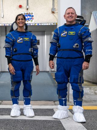 two astronauts standing side by side on a tarmac in spacesuits. white covers protect their boots