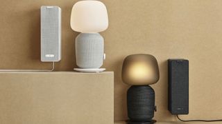 Two Sonos IKEA Symfonisk table lamp speakers (one white, one black) next to each other on a beige surface in front of a beige wall. The white speaker is raised higher than the black.