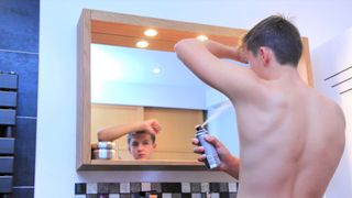 young teen boy using a spray deoderant on his left armpit while he looks in a bathroom mirror