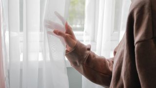 A person moves a thin, see-through curtain aside to look out through the window behind.