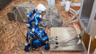 a robot on wheels with head and long arms and hands. the robot's hand is stretched towards a tube. the tube is on top of a metal platform. in the background is a simulated rock, sand and struts indicating a practice mars base