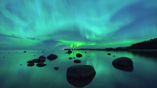 A blue-green sky with streaks of auroras over a body of water with rocky silhouettes.