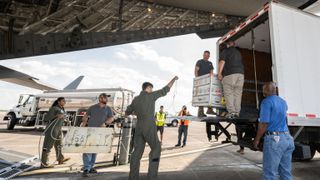 workers unload a silver crate from a plane and put it in the back of a truck.