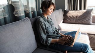 A woman sitting on a couch and smiling while working on a laptop