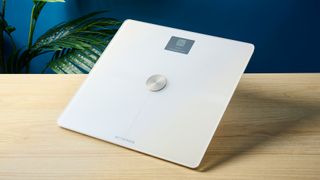 a white smart scale by withings rests on a wooden table, showing its LED screen and weighing scales