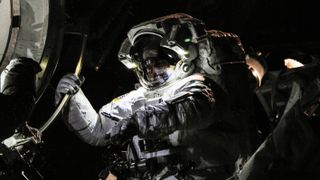 a ghostly lit picture of astronaut chris cassidy in a spacesuit in space. the astronaut holds on to the international space station hatch with his right hand