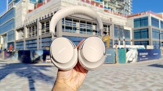 Listing image for best noise-cancelling headphones showing Bose QC Ultra Headphones listing image shown in smoke white held aloft in daylight on an urban street