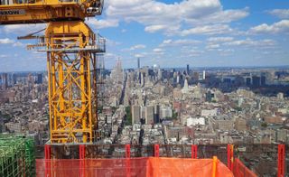 Daytime image from the roof of a high rise, looking across the skyline, lower buildings and high-rise towers in the distance, yellow crane on the roof at the forefront,, cloudy blue sky