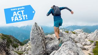 A trail runner dressed in blue cresting a mountain peak with other distance mountains in the background against a white cloudy sky.