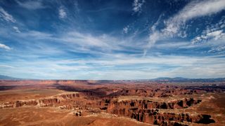 red rock structures and canyons below a blue sky with wispy white clouds.