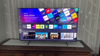 The Hisense 55E7K Pro TV on a TV stand in front of a wall covered in tree wallpaper. On screen is the VIDAA App Store showing various video streaming apps like Netflix, BBC iPlayer, ITVX and Apple TV+.