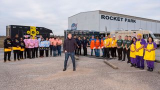 a man in jeans and a jacket stands in a parking lot with about 20 people wearing multicolored aprons surrounding him