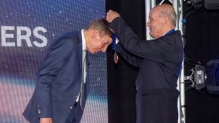 an older man drapes a medal around the neck of a man on a stage