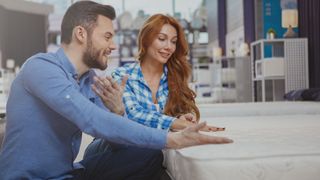 A man with dark hair and a woman with red hair go mattress shopping together