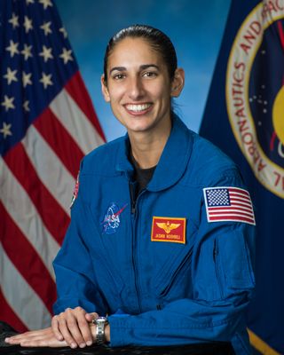 A woman sits in front of an american flag and nasa flag, wearing a blue jumpsuit.