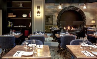 A restaurant with decorated dining tables, a patterned rug, a round wall mirror and wall shelving.