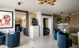 Alternative view of the seating area at Bar Roc at Ellerman House featuring blue and green seating, stone and white walls, multiple ceiling lights, grey and bronze coloured round tables, wall art, a small elephant statue on a plinth and green plants in pots. There is also a partial view of the bar area