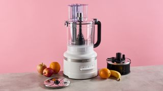 A white colored KitchenAid 13 cup food processor photographed against a pink background, sitting on a great stone-effect surface. The food processor is surrounded by apples, oranges and a banana. The accessory storage caddy is pictured to the right of the processor, and the reversible grating disc is on the left.